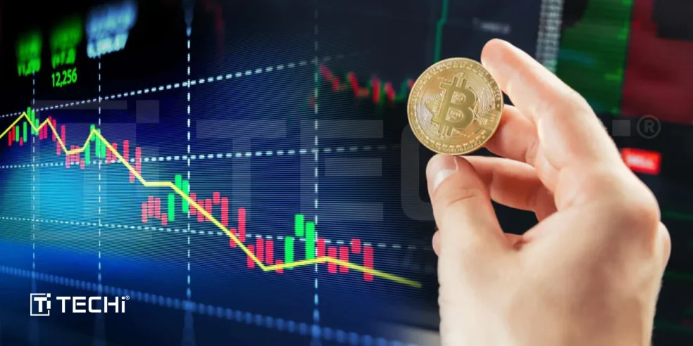 A hand holding a Bitcoin coin in front of a digital stock market chart showing a price decline.