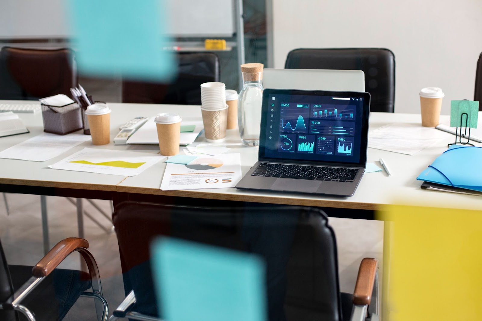 A laptop displaying data analytics dashboards is placed on a meeting room table surrounded by coffee cups, documents, charts, and office supplies.