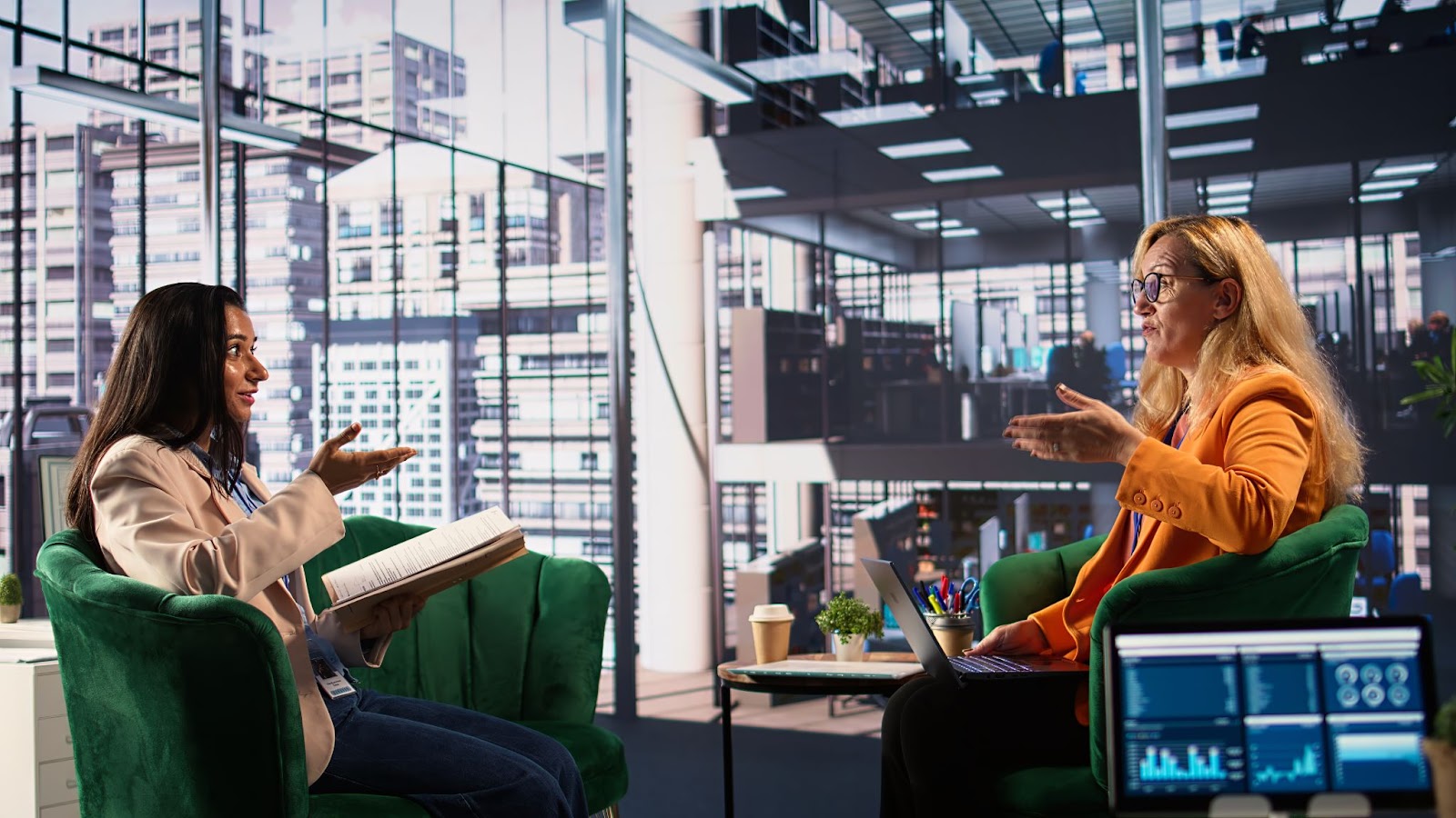 Two women are seated in green armchairs in a modern, glass-walled office space. One woman, dressed in a light blazer and jeans, is holding a large open notebook and gesturing as she speaks. The other woman, dressed in an orange blazer, is sitting opposite her with a laptop on her lap and a coffee cup and plant on the side table. Office backgrounds filled with desks, computers, and people are visible through the large windows. A third screen with data charts is partially visible in the foreground.